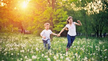 Little boy aged 5 and his elder sister aged 9 are running on dandelion field. They are enjoying beautiful nature and the sunset, laughing happily. Focus on the little boy, the girl is slightly soft.