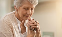 Senior woman praying while holding christian symbol of crucifix. Old woman praying to god with hope and closed eyes. Elderly believer make a prayer with faith holding rosary in hands.